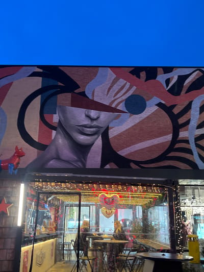 This image captures a segment of a commercial building and an adjacent cafe storefront in Reichstett, France. The upper approximately one-fifth of the frame displays a clear, uninterrupted deep blue sky.

Immediately below the sky, a large mural is painted onto a light brown brick wall. The mural depicts the lower half of a stylized female face, from the nose bridge to the neck, rendered in shades of grey and purple, with visible lips and chin. The upper portion of the face, where the eyes and forehead would be, is obscured by a prominent, dark red triangular shape, which is integrated into an abstract composition of various geometric and organic forms in colors including beige, light blue, black, and additional shades of red. To the left of the mural, at the top edge of the structure below, a small, three-dimensional decorative object resembling a red and blue equine figure is present.

Occupying the lower half of the image is the transparent, enclosed facade of a restaurant or cafe. This area is defined by clear panels, possibly glass or plastic, and features numerous warm-toned string lights draped along its interior upper edge. Inside this illuminated enclosure, multiple dark, round tables with corresponding chairs are visible. Two adult women are seated at one of these tables, positioned centrally within the cafe area; one faces left, the other faces right, suggesting an interaction. Suspended from the ceiling within the cafe are two heart-shaped balloons: one predominantly red and white, the other metallic gold or silver. To the far left of the cafe's transparent section, a vertical, white LED strip light is affixed to a dark, textured wall panel that also displays a red star ornament. Reflections on the transparent facade reveal additional interior elements and subtle indications of an exterior environment, including a yellow outdoor waste bin and a mannequin dressed in a brown garment visible through or reflected on the far right panel.