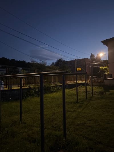 The image shows an outdoor scene at night in the city of Troyan, Bulgaria. A set of parallel bars are located in a grassy area next to a fenced structure. A streetlight illuminates the scene, casting a bright glow on the grass. Overhead, power lines stretch across a dark blue sky. A building is visible in the background. The perspective is from a low angle, with the camera tilted to the side.