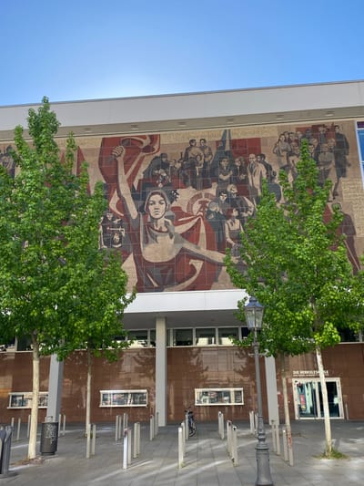 A daytime shot shows a building facade with a large mural in Dresden, Germany. The mural, constructed of brick or tile, depicts people in a revolutionary or socialist realist style. The main figure appears to be a woman holding what might be a fan or flag. Other figures in the mural are soldiers and civilians. In front of the building, there are young trees and a street lamp with a lamphouse. There are metal bike racks and a litter bin on the pavement. The building's architecture features rows of windows and tiled panels. The building appears to be the “Die Herkuleskeule”, a cabaret theater in Dresden.