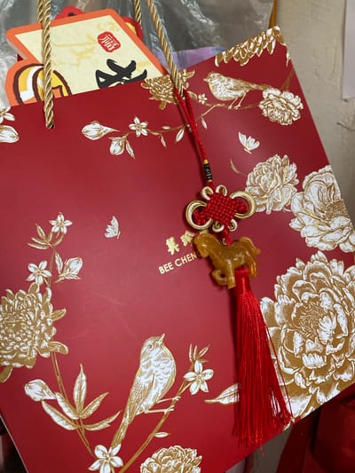 The image displays a close-up view of a red gift bag adorned with gold foil floral and bird motifs. A decorative tassel featuring a horse charm hangs from the top of the bag. Partially visible inside the bag are what appear to be greeting cards with Chinese characters and red stamps. The text "BEE CHEN" is printed on the gift bag. The scene is a static indoor shot with no discernible city or environmental context.