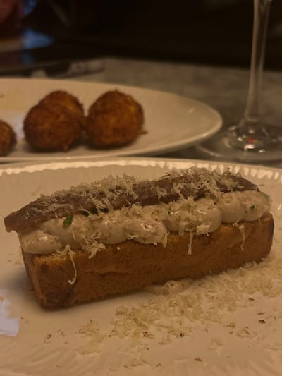 The scene depicts a close-up of a meal served indoors at an establishment in Santander, Spain. In the foreground, a rectangular piece of toasted bread, possibly a pintxo, is centered on a white plate with a ridged edge. This toast is topped with a generous layer of light-colored, creamy paste or mousse, adorned with several dark brown anchovy fillets and finely grated white cheese. Scattered flakes of the grated cheese are also present on the white plate surface around the toast. In the blurred background, another white, oval-shaped plate holds at least two spherical, golden-brown, deep-fried items, consistent with croquettes. To the far right, a partial view of a clear wine glass is visible. The environment is dimly lit, suggesting a restaurant or bar setting. No individuals are visible.