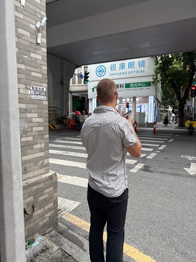 The image shows a street scene in Yuexiu District, China, under an overpass. In the foreground, a man is standing facing away from the camera, looking at his mobile phone. He is wearing a patterned short-sleeved shirt and black pants. Behind him, a pedestrian crossing is visible, with a traffic light showing green in the distance. A building with signage that reads "SHI KANG OPTICAL" is also present. Further back, several people appear to be sitting or standing near what looks like a makeshift stall or collection of belongings. The overall environment suggests a busy urban area.