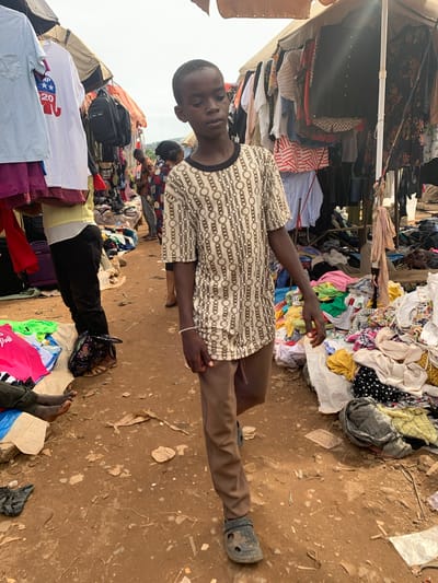In Nyanya, Nigeria, a person is captured in a medium shot, lying down amidst a bustling market scene. They are wearing a light-colored patterned shirt and brown trousers, with a black shoe on their right foot. The person's face is partially visible, with their eyes closed or looking away, giving an impression of rest or perhaps fatigue.
The background is filled with various clothing items, suggesting a second-hand clothing market. Piles of clothes are stacked on shelves and on the ground, showcasing a variety of colors and patterns. Some items are neatly folded, while others appear to be in disarray. In the distance, more market stalls and people can be seen, indicating a busy and active environment.
The ground is dusty and unpaved, typical of outdoor markets in the region. The lighting suggests it is daytime, though the exact time is difficult to ascertain. There are no prominent weather cues, but the overall atmosphere feels warm and perhaps humid.
One notable detail is a white t-shirt with a red elephant logo and the text "TRUMP 2020" printed on it, partially visible in the lower left of the image, tucked under other clothes. This political merchandise adds an unexpected element to the otherwise ordinary market scene. The scene conveys a sense of everyday life in a Nigerian market, with the individual's relaxed pose offering a moment of calm within the energetic surroundings.