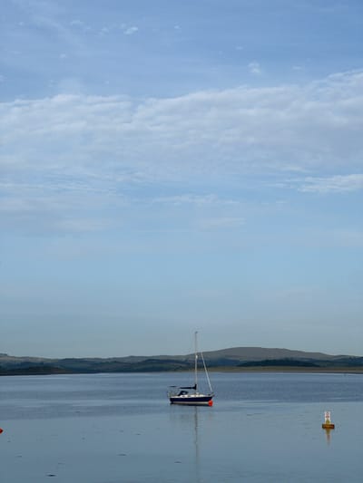 This media file captures a serene waterscape. The scene is dominated by a vast expanse of calm water, reflecting the pale blue sky and scattered clouds. On the right side of the frame, a lone sailboat, predominantly white with blue accents, is moored. Its mast stands tall, reaching towards the sky, and a small orange buoy is visible near its stern. Further in the distance, another buoy with a warning symbol, a red triangle with an exclamation mark, floats on the water.

The background features a gentle, rolling landscape of hills and coastline, shrouded in a soft, hazy light. The time of day appears to be late afternoon or early evening, given the diffused light and the cool, muted tones of the scene. The weather is overcast but tranquil, with no indication of harsh conditions. The overall mood conveyed is one of peacefulness and solitude.

No people are visible in the frame. The image appears to be a landscape photograph, likely taken in or around Terhillion, Ireland, given the subtle hints of the Irish coastline. The visible text on the buoy is "CAUTION," enclosed within a red triangular warning symbol.