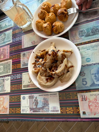 The image shows a table covered with various currency bills from different countries, including Russia, Canada, Singapore, United States, United Kingdom, and Myanmar. On the table, there are two plates of food. One plate contains fried dumplings, and the other contains steamed dumplings drizzled with a dark sauce. A glass pitcher filled with a light-colored liquid and some floating ingredients is also visible. A hand is holding a fork and picking up one of the fried dumplings. The table is adorned with a vibrantly patterned textile. The background is slightly out of focus, but it suggests an indoor setting. The image does not provide any specific visual cues that would allow for the identification of the location as Bhagsu, India.