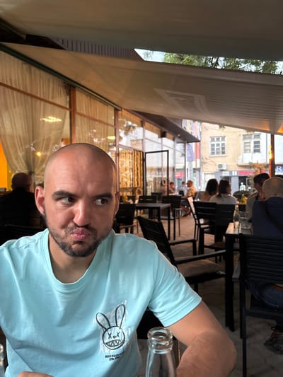 An outdoor cafe scene in Karlovo, Bulgaria, is depicted. In the foreground, a bald adult male with a short beard is seated, wearing a light blue t-shirt featuring a stylized bunny graphic with crossed eyes and sharp teeth, and the text "BREAK RULES CLUB ESTD. 1992." He is looking to his left (viewer's right) with pursed lips, exhibiting a distinct facial expression. A clear glass bottle is partially visible on the table in the lower left.
The midground shows an active cafe environment with multiple patrons, most of whom are blurred, seated at dark tables with matching chairs. A person wearing a dark shirt is partially visible to the primary subject's right (viewer's left). Behind the seated individuals, a building with large windows and light-colored interior curtains suggests an indoor section of the establishment or an adjacent building.
The background reveals an urban street with older buildings. One building facade displays fragmented commercial signage including "JEANS WEAR" and "PAL...". Graffiti or street art is visible on a wall in the upper left portion of the background. The outdoor seating area is covered by a light-colored, possibly retractable, awning. Above the awning, a narrow strip of trees and clear sky is visible.
