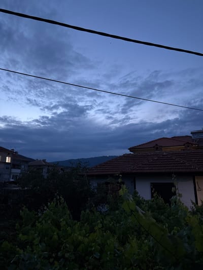 This is a vertical, wide-angle, eye-level shot of a cloudy sky at dusk over a residential area in Strelcha, Bulgaria. The sky is a deep blue, transitioning to lighter shades near the horizon, with dramatic, wispy clouds stretching across. A dark, thick electrical wire runs diagonally from the bottom left corner towards the top center of the frame, with a thinner wire visible on the right side.
In the middle ground, the terracotta tiled roofs of several buildings are visible, one with prominent dormer windows. A white building with dark windows is nestled beside it. To the right and in the foreground, lush green foliage, likely from a vine or tree, dominates the frame, partially obscuring the buildings. The leaves are dense and varied in shade due to the low light.
Further down the hill, another building is partially visible with illuminated windows, suggesting nighttime is approaching. The overall mood is subdued and peaceful, enhanced by the twilight atmosphere and the soft glow from the building's lights. The scene depicts a quiet evening in a town where nature and infrastructure coexist.