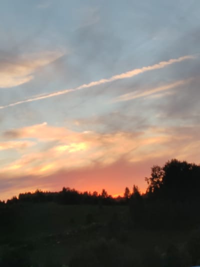 This is a photograph of a sunset over a rural landscape in Chukhlem, Russia. The sky is filled with dramatic clouds, painted in hues of orange, pink, and pale blue, with streaks of white contrails crisscrossing the upper atmosphere. Below the vibrant sky, a dark silhouette of trees and rolling hills stretches across the horizon. The foreground is also dark and indistinct, suggesting it is evening. The overall mood is peaceful and serene, capturing the beauty of a natural sunset. No people, objects, or text are visible in the image.