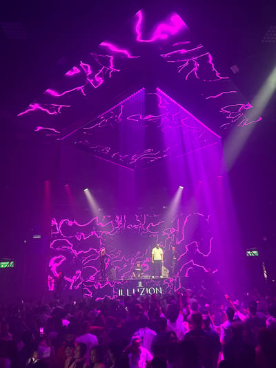 This is a vertical shot of a lively concert or club scene at night, likely in Patong, Thailand. The dominant color is a vibrant purple, emanating from stage lighting and LED screens that display abstract, neon-like pink graphics. A large crowd of people, densely packed, fills the right side of the frame, their faces illuminated by the purple glow. Many are raising their hands, suggesting excitement and engagement with the performance.
On a stage to the left, a performer is visible, dressed in dark clothing. There are also DJ decks and sound equipment. The stage itself is adorned with the same glowing pink graphics as the large screens. Several beams of white and purple light cut through the smoky atmosphere, adding to the dynamic ambiance.
The image captures a moment of high energy and celebration, typical of a nightlife event. The presence of "EMERGENCY EXIT" signs suggests an indoor venue. The crowd's poses and facial expressions indicate a positive and enthusiastic response to the music and visual spectacle. The time of day is clearly night, given the artificial lighting and the context of a concert or club.