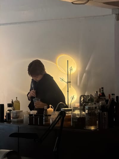 The image depicts a dimly lit interior, likely a bar or cafe, with a person dressed in black working behind a counter. The person is holding a clear plastic cup and appears to be mixing something with a utensil. Behind them, a wall is illuminated by a decorative lamp that casts a soft, warm glow, creating circular patterns of light. The counter is lined with various containers, including glass jars filled with what appear to be dried ingredients, bottles, and dispensers. To the right, several bottles, some with dark liquid, are arranged on the counter. The overall ambiance is intimate and possibly nocturnal, suggested by the low lighting and the presence of bottles that might contain alcoholic beverages. The scene is devoid of any specific identifying landmarks that would definitively place it within Chiang Mai City Municipality, Thailand, but the style of the bar and the arrangement of goods could be consistent with establishments found in such a location.