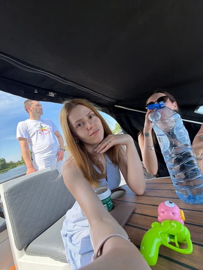 In Gellicum, Netherlands, a group of three individuals are on a boat on a sunny day. A man in a white t-shirt and shorts stands in the background, looking to his left. In the foreground, two women are seated. The woman on the right holds a large bottle of water, obscuring her face with it. The woman on the left is closer to the camera, holding a coffee cup and resting her chin on her hand. A green water gun and a pink octopus-shaped toy are on the wooden table in front of them. The boat has a black canopy overhead and grey seating. The sky is blue with some scattered clouds.