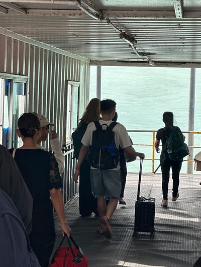 The image captures a group of people disembarking from a ferry onto a walkway overlooking the sea. The location is identified as Nathon, Thailand, a busy ferry terminal. The walkway appears to be made of corrugated metal, with a corrugated metal roof overhead supported by beams. The walls are also corrugated metal, with small windows on the left side. Sunlight streams in from the right, creating strong shadows on the walkway.
In the foreground, a woman in a black top with lace sleeves is holding a red duffel bag. To her left, another person, partially visible, is wearing a dark blue jacket. Behind them, a man with short hair and a backpack is pulling a black rolling suitcase. He is wearing a white t-shirt and denim shorts. To his right, a woman with long blonde hair is walking ahead. Further to the right, a person wearing a black t-shirt and dark pants is carrying a green backpack. They are all moving in the same direction, away from the ferry. The sea is visible in the background, with gentle waves. The overall scene depicts the common activity of ferry travel, with passengers disembarking with their luggage.