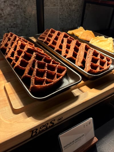 The image shows a close-up view of chocolate waffles and some thin, golden crepes arranged on dark metal trays. The waffles are stacked in two trays, with a few crepes visible in the upper left tray. The waffles have a deep brown color and a distinct grid pattern. The scene is set on a wooden surface with a carved inscription that appears to read "Blog S". A small white card with a clear plastic cover is placed on the wooden surface. Visible text on the card reads "Waffles de Chocolate" and "Chocolate Waffle". The lighting suggests it is daytime, with strong shadows cast across the wooden surface, highlighting the texture of the waffles and wood. The overall setting appears to be a food display or serving area, possibly in a cafe or restaurant.