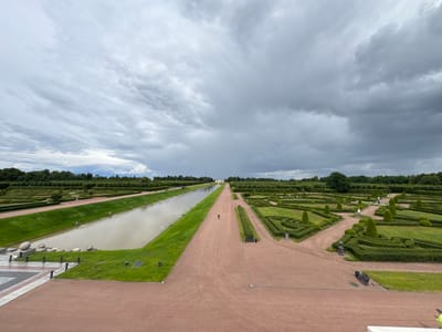 The image depicts a formal garden in Strelna, Russia, likely part of the Konstantinovsky Palace complex, under a dramatic, cloudy sky. The scene is characterized by symmetry and manicured landscapes.
In the foreground, a wide, reddish-brown pathway dominates, leading towards a long, rectangular canal on the left. This canal is bordered by a neatly mown green lawn and the pathway. Along the edge of the lawn, several large, pale, oval-shaped stones are arranged, with low, dark bollards and chains in front of them. Further to the left, a section of patterned paving can be seen.
The canal stretches into the distance, with a lone figure walking on a path alongside it. The water in the canal is a murky, brownish-grey color. Beyond the canal, the landscape unfolds into a series of meticulously shaped hedges, forming intricate geometric patterns. These hedges are interspersed with areas of green grass and smaller trees. In the far distance, a row of dense green trees forms the horizon, and a pale yellow building is visible through a gap in the trees.
The sky is filled with heavy, dark gray clouds, suggesting an overcast or approaching storm. The overall impression is one of grand, formal landscaping contrasted with the dynamic, moody sky.