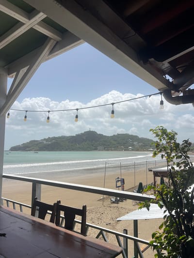 This is a photograph taken from a covered outdoor seating area overlooking a beach in San Juan del Sur, Nicaragua. The viewpoint is from a wooden table and chairs on a balcony, looking out towards the ocean.
In the foreground, a portion of a wooden table is visible, with two dark wooden chairs behind it. A white railing separates the balcony from the beach area.
The beach is sandy and appears to be mostly empty, with some beach chairs and umbrellas set up in the distance. There are some structures on the beach, including a white-roofed cabana. String lights are hung across the view, adding a decorative element.
The ocean is a calm turquoise color, with gentle waves breaking on the shore. In the background, a lush green hill rises from the coastline, dotted with buildings and homes. At the very top of the hill, a prominent statue of Jesus Christ can be seen, a landmark of San Juan del Sur. Several boats are anchored in the bay.
The sky is a bright blue with fluffy white clouds. The overall impression is a sunny, relaxed atmosphere, characteristic of a tropical beach town. The photograph captures the iconic view of the Cristo de la Misericordia statue overlooking the bay.