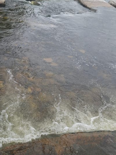 The image shows a close-up, top-down view of a shallow body of water flowing over rocks. The water is mostly clear, with some darker areas indicating deeper sections or submerged rocks. The surface of the water is disturbed, creating ripples and small white patches of foam, particularly on the right side of the image where the water appears to be moving faster. The rocks beneath the water are varied in color, ranging from light grey to reddish-brown, and some have a smooth, worn appearance. On the left side of the frame, larger, lighter-colored rocks are visible, partially submerged. The overall impression is of a natural, outdoor scene, likely a stream or river. The lighting suggests it could be daytime, with no strong shadows indicating direct sunlight. There are no people or man-made objects visible. The scene evokes a sense of natural movement and the textures of water and stone.