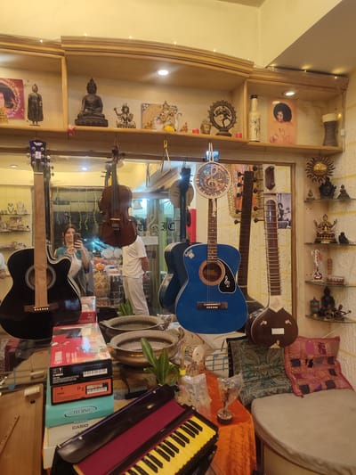This image captures the interior of a shop in Puttaparthi, India, specializing in musical instruments and spiritual artifacts. The scene is dominated by a large mirror reflecting much of the store and a woman taking a photo with her phone. She is smiling and wearing a blue and grey patterned garment. Another person, partially visible in the reflection, wears white clothing.
Numerous musical instruments are on display. In the foreground, a black acoustic guitar stands upright on the left. A bright blue acoustic guitar, possibly branded "GIBSON" (partially visible), hangs near the center, decorated with a circular motif featuring a musical note. To its right, a dark brown sitar is prominently displayed. Hanging above are a dark wooden string instrument resembling a cello or violin, and another string instrument. In the foreground on a table, a yellow and black keyboard (harmonium) is visible, along with several boxes for electronic keyboards, including "Yamaha PSR-A50," "TECHNO," and "CASIO SA-46."
The shop's shelves are laden with an array of spiritual and decorative items. These include various statues of Buddha in different poses, figures of Hindu deities such as Shiva Nataraja and what appears to be Ganesha, and framed images of spiritual leaders like Sai Baba. Other decorative elements include bottles, small pots, and a stylized sun motif on the wall. Small fairy lights adorn the top of the shelving units. Colorful, embroidered cushions are arranged on a low seating area on the right.
The overall setting suggests a vibrant retail space, likely catering to tourists or devotees in Puttaparthi, offering both musical instruments and items of spiritual significance. The woman's smile indicates a pleasant shopping experience. The visible text on the guitar headstock reads "Twelfth note," and a reversed "DESIGN" sign is visible in the mirror. The artificial lighting suggests it could be any time of day, but the indoor setting is the primary focus.