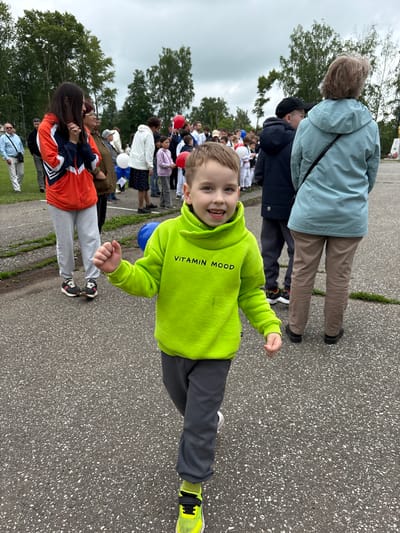 An outdoor scene in Kuyeda, Russia, features a young boy in the immediate foreground, centered and walking towards the camera. The boy, approximately 5-7 years old, has light brown hair, blue eyes, and an open-mouthed smile. He wears a bright lime-green hooded sweatshirt with "VITAMIN MOOD" printed in black, dark grey sweatpants, and black shoes with neon yellow accents and socks. His fists are loosely clenched.
Behind the boy, an assembly of individuals, primarily adults and children, occupies the midground and background. To the left, a woman with long dark hair, wearing an orange, white, and navy blue athletic jacket and light grey sweatpants, stands with clasped hands. Behind her, a man in a light blue shirt and sunglasses walks. To the right, the back of a woman in a light blue hooded jacket and khaki pants is visible, along with a boy in a black cap and dark jacket.
Further back, a crowd includes children dressed in white martial arts uniforms (gis), suggesting a sports event or demonstration. Red, white, and a partially visible blue balloon are interspersed among the attendees. The ground consists of dark asphalt with white aggregate, bordered by green grass and numerous trees in the background. An overcast sky is visible above. On the far right background, a stone monument with a golden element on top is discernible.