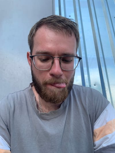A young man with a beard and glasses is sticking his tongue out at the camera. He is wearing a gray t-shirt with orange and white stripes on the sleeves. The background is a blue and white corrugated metal wall. The photo appears to have been taken outdoors, possibly in an urban setting in Pivdennoukrainsk, Ukraine.
