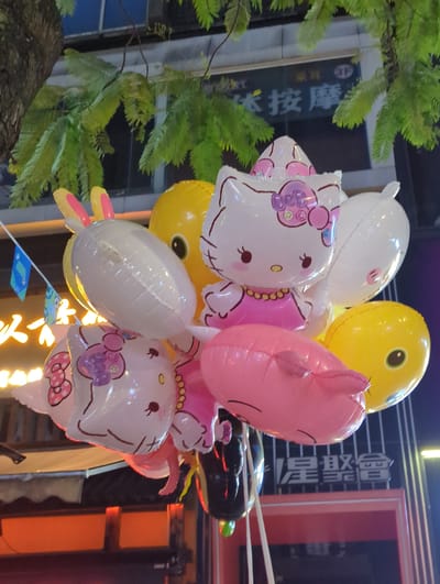 The image shows a cluster of helium balloons hanging from tree branches at night. The balloons are colorful and feature cartoon characters like Hello Kitty and yellow ducks. In the background, there is a building with Chinese characters and neon lights. The overall scene appears to be a street market or a festive outdoor event in Longta, China. There are no people visible in the image.