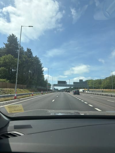 The image shows a highway in Elmbridge, United Kingdom, viewed from inside a vehicle. The road has multiple lanes with white lane markers. There are other cars on the road. Highway signs are visible above the road. Trees line the side of the highway. A barrier is visible along the side of the road with orange cones. The sky is blue with clouds. The interior of the vehicle is visible in the foreground.