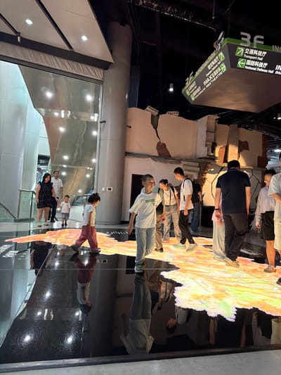 The image depicts an indoor scene with several people, including adults and children, walking across a large, illuminated floor display. The floor display appears to be a map or a geographic representation with vibrant colors and textures. Some individuals are walking on the display, while others are standing nearby. In the background, there is a structure that resembles a partially collapsed building, with exposed brick and cracked walls, suggesting a display related to historical events or disaster simulation. Overhead, signage is visible, with one sign indicating "3F INFORMATION" and listing various halls such as "Transport Hall," "Disasters Defense Hall," "National Defense Hall," and "IMAX THEATRE," with corresponding Chinese text. The environment is modern, with polished floors reflecting the lights and the illuminated display, and large pillars and metallic surfaces suggesting an exhibition hall or museum setting within 江北城街道, China.