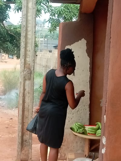 The image displays a scene in Nkwelle Ezunaka, Nigeria. A woman is positioned with her back partially to the viewer, facing slightly right. She wears a sleeveless black dress and has dark braided hair styled in a bun. A beaded earring hangs from her visible left ear. She holds a small, red rectangular object in her right hand and a black plastic bag in her left hand. To her right, a metal tray on a wooden stand contains multiple bunches of unripe green bananas. The wall directly behind the bananas is reddish-brown, featuring a prominent patch of lighter, exposed plaster. A rough, grey concrete pillar stands vertically to the woman's left. The ground in the foreground is reddish-brown dirt. In the background, beyond the pillar, an open dirt area with sparse green vegetation is visible, from which a plume of light-colored smoke rises. Further back, numerous green trees and several distant residential buildings with light-colored walls and dark roofs are discernible on elevated terrain. Partial construction blocks are also visible in the mid-background, and the faint outline of a vehicle is present on a distant dirt road to the far left.