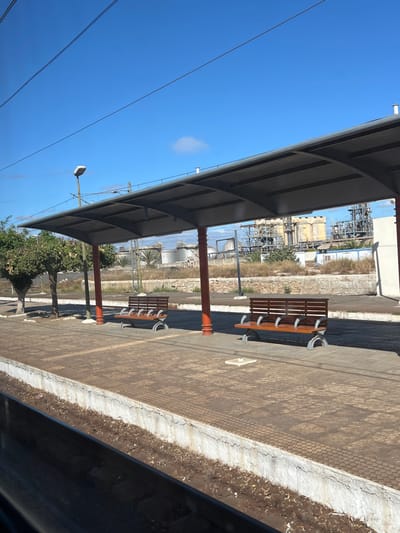 The image captures a static, uninhabited train station platform in Sidi Kacem, Morocco, under a clear, bright blue sky containing a single wispy cloud. The immediate foreground shows the dark, blurry edge of a viewing window or train carriage, partially obscuring the railway tracks and a dirt-laden concrete platform edge exhibiting minor cracks. The platform surface is paved with light brown or grey concrete tiles, including tactile paving elements along the edge.

Mid-ground details include a grey, curved metal canopy supported by several reddish-orange pillars, providing shelter over the platform. Beneath this canopy, two empty wooden benches with grey metal frames are positioned facing the tracks. To the left, a light pole with a grey luminaire is visible, along with a few small trees featuring green foliage behind the platform.

The background reveals a dry, unkempt area with sparse vegetation and bare earth, separated by a low stone wall. Beyond this, a prominent industrial complex is situated, characterized by numerous large, metallic storage tanks (both cylindrical and spherical), extensive networks of pipes, and scaffolding-like structures, indicative of a refinery or processing plant. Several taller industrial buildings are integrated within the complex. Overhead, multiple thin power lines or catenary wires stretch diagonally across the sky.