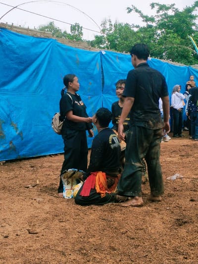 The scene takes place in a dirt lot with a large blue tarp behind the subjects. The tarp is attached to a wooden structure. The tarp is made of a sturdy, thick material. The subjects are standing and sitting in front of the tarp. There are trees and foliage beyond the tarp. The ground is brown and covered in dry leaves, grass, and other debris. There are six people in the scene. Three are men and three are women. One of the men is facing the camera. He is wearing a black shirt, black pants, and black shoes. He has short dark brown hair and is wearing a watch on his left wrist. His hands are on his hips.  One of the women is facing the camera. She is wearing a black shirt, black pants, and black shoes. She has long dark brown hair and is wearing a black and red scarf. She is carrying a white and brown bag on her right shoulder. Her arms are at her sides and her hands are closed. The woman is looking at the camera. The remaining subjects are facing away from the camera.  The subject to the left is wearing a black shirt, black pants, and black shoes. The subject on the right is wearing a black shirt, black pants, and black shoes. The two men standing on the right are wearing black shirts, black pants, and black shoes. One of the women is wearing a light blue shirt, blue pants, and a long white veil. One of the women is wearing a black hijab, grey pants, and a light blue shirt with a design on it. The man in the black shirt on the right is looking at the subjects in front of the tarp, and the other men are looking to the left. The women standing on the right are looking to the left. The man in the black shirt is standing and the other subjects are sitting on the ground. The man in the black shirt is looking at the camera. The subjects on the ground are looking down.  The subjects are standing and sitting in a relaxed, casual manner. The subjects appear to be waiting for something or someone. The subjects are in a location in Kecamatan Kedawung, Indonesia. The location's coordinates are latitude -7.494, longitude 111.07.