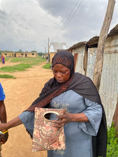 The image depicts an outdoor scene in what appears to be a semi-urban or rural setting. The primary subject is a woman, likely of middle age or older, who is looking slightly towards the viewer with a gentle, soft smile. She wears a black headscarf (hijab) adorned with intricate orange beaded patterns, and a light blue or grey long-sleeved dress featuring white or silver embellished designs on the chest.

The woman is holding a decorative clutch bag or purse with a metallic, possibly silver-colored, handle. The bag has a distinct paisley-like pattern in shades of cream, red, and brown. In the foreground, part of another person's arm and a blue shirt are visible, seemingly in the process of interacting with the woman's hand or the object she is holding; a yellow band is on the other person's wrist.

The immediate background includes simple structures made of corrugated metal sheets and rough wooden beams, suggesting a utilitarian or makeshift building. Beyond this, a wide, open area of dusty brown earth mixed with patches of green grass stretches into the distance, resembling a field or unpaved public space. In the far background, a long, low building with multiple windows is visible, possibly a school or community center, with scattered smaller figures and what appear to be motorcycles or other vehicles. Power lines cross the sky.

The sky is overcast with heavy clouds, indicating it is daytime but without direct sunlight. The overall atmosphere appears casual and everyday. No discernible text is visible in the image. The setting and building styles are consistent with a location in Riyom, Nigeria.