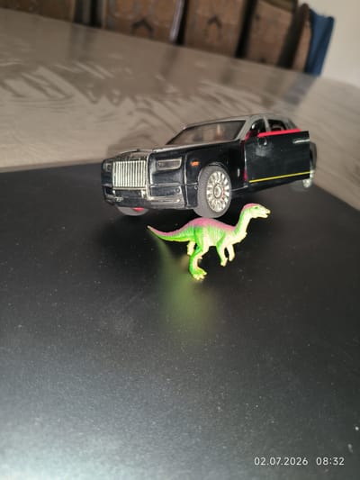 The image depicts a toy black Rolls-Royce car with a red-lined open door and a green and pink toy dinosaur on a black surface. The background shows a blurred scene with wooden structures and a reflective surface. The image has a timestamp in the lower right corner: "02.07.2026 08:32". There is no indication of the city of Kattasu, Uzbekistan, within the image.