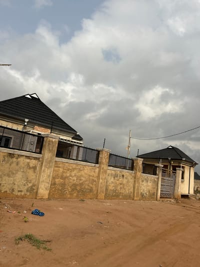 The scene depicts an exterior view in Ilishan, Nigeria, under an overcast sky. In the midground, a textured, light brown concrete perimeter wall with prominent vertical pillars runs horizontally. This wall is surmounted by a dark metal fence composed of horizontal slats, fitted with electric fence insulators and wires.
Behind the wall to the left, a portion of a two-story building is visible. This structure features a dark, possibly charcoal-gray, tiled hip roof with white trim, and a light-colored upper facade with dark horizontal window grates. An area of exposed red brickwork is also present on its facade. To the right of this, also situated behind the wall, stands a smaller, single-story, light-colored octagonal building with a matching dark tiled hip roof. Its facade incorporates a gray metal entrance gate with horizontal slats and a visible window opening to the right of the gate. A slender wooden utility pole with an overhead electrical wire is positioned behind the wall, slightly right of center.
The foreground consists of reddish-brown dirt soil with scattered patches of sparse green vegetation. A blue and white patterned fabric item, likely an article of clothing, and fragments of white and red plastic debris are visible on the dirt surface in the lower left portion of the image. The upper portion of the image is dominated by a sky heavily covered with dense gray and white clouds, with a small area of lighter blue visible through a gap in the upper-left. No individuals are present in the scene.