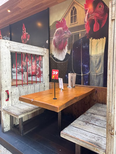 The image captures an interior scene, likely a restaurant in Costa Mesa, United States of America. The focus is on a table with a wooden surface, holding a napkin dispenser and a table number (39) in a red holder. The backdrop is a collage featuring images of chickens in various scenes, including one wearing overalls and holding a pitchfork against a cosmic backdrop, and others within a chicken coop. The furniture appears rustic, with aged or distressed wood finishes. The location seems to be a unique establishment, blending elements of farmhouse decor with whimsical chicken-themed art. The overall atmosphere suggests a casual, possibly farm-to-table dining experience.