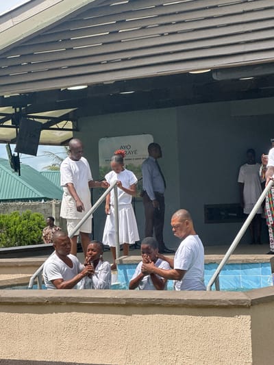 In Itoga, Nigeria, a baptismal ceremony is taking place in an outdoor setting under a covered structure. The water in the baptismal pool is turquoise and appears to be about waist-deep for those in it. Several individuals are involved in the ceremony, all dressed in white. In the foreground, a man is baptizing a woman in the pool, with his hands on her head. To the left of this pair, another woman is also being baptized by a man. Further back, a man in a white t-shirt and shorts, presumably a religious leader, is standing next to a young girl in a white dress, holding her hand as she enters the pool. Behind them, other figures are visible, including a man in a blue shirt and brown pants, and a woman in a white t-shirt and shorts. A large sign with text is visible on the wall in the background, and part of a building with green roofing is seen to the left. The overall atmosphere suggests a religious gathering focused on the sacrament of baptism.