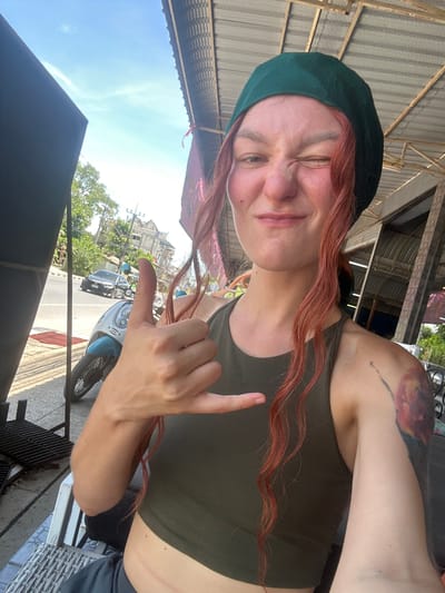 A woman with red hair and a green beret is taking a selfie on a street in Nathon, Thailand. She is wearing a green tank top and has a tattoo on her right arm. She is making a "shaka" hand gesture. In the background, there are buildings, trees, a road with a car and a motorbike, and a corrugated metal roof overhead. The sun is shining.