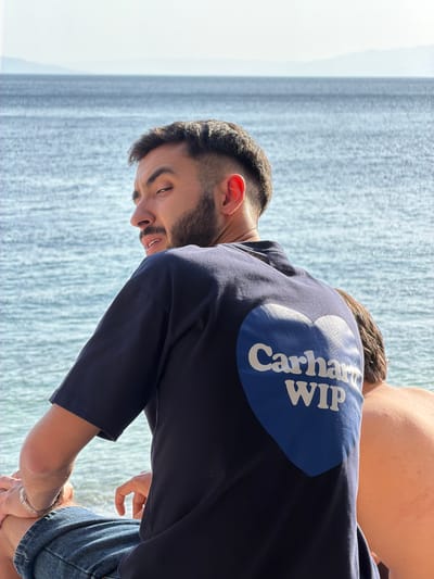 The image, captured in Grad Rijeka, Croatia, depicts two individuals outdoors on a clear, sunny day. The prominent subject is a man with a beard and dark hair, seen from behind and side. He is leaning forward, looking downwards and to his left with a focused or slightly strained expression, his mouth agape. He wears a dark navy blue t-shirt featuring the brand "Carhartt WIP" in white letters within a distinctive blue circular logo on its back. A silver-toned watch or bracelet adorns his right wrist. Behind him, another person is partially visible, also bending over, with their back towards the camera. The background consists of a vast, calm body of water, likely the Adriatic Sea, showing subtle ripples and reflecting the bright sunlight. The overall scene conveys a relaxed outdoor setting by the water.