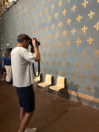A man is taking a picture in a room with a ceiling adorned with golden fleur-de-lis symbols on a blue background. The room has a tiled floor, and some modern chairs are visible. Other people are in the background. The location is likely in Florence, Italy, given the architectural style and fleur-de-lis imagery.
