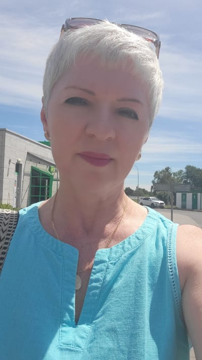 This is a portrait of a woman with short, light-colored hair, wearing a light blue top and a gold necklace. She is outdoors, with a bright blue sky and scattered white clouds in the background. A building with a green-tinted window is visible behind her. The sun is shining, suggesting it is daytime. The woman's face is turned towards the camera, and she has makeup on her lips. Sunglasses are pushed up onto her hair. The scene appears to be in Belaazyorsk, Belarus.