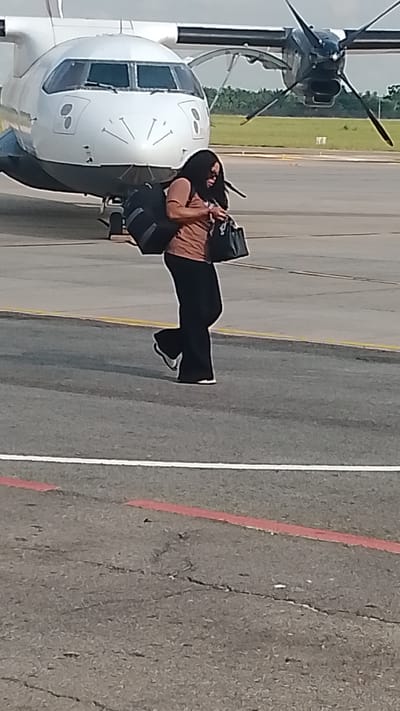 A full-body, medium-shot photograph taken outdoors shows a woman walking on an airport tarmac near a turboprop airplane. The woman, who has long black hair, is wearing a rust-colored short-sleeved top and black flared pants. She is carrying a black tote bag in her right hand and has a large dark duffel bag slung over her left shoulder. Her white sneakers are visible. The airplane, a white turboprop with dark-colored propellers, is parked behind her, and its cockpit windows are visible, giving it a face-like appearance with lines resembling a smile. The tarmac is gray asphalt, marked with white and red lines. The background shows a grassy area with some trees and a hazy sky. The scene is likely at an airport in Orerokpe, Nigeria, given the context.