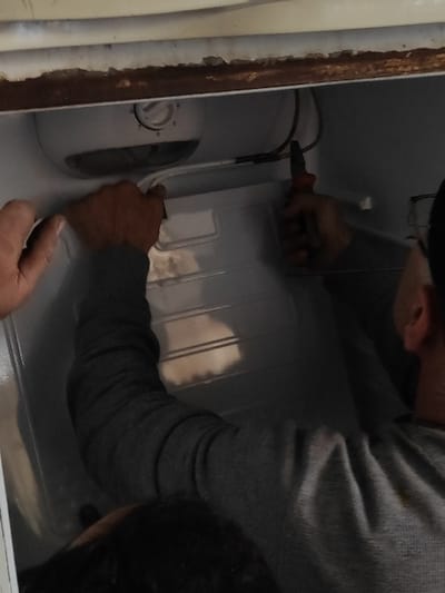 In Ash-Shaghour Municipality, Syria, two individuals are engaged in maintenance work on the interior of a white refrigerator. One person, wearing a grey long-sleeved shirt, is positioned with their right arm extended into the appliance, using red-handled pliers to manipulate a wire or tube. The left hand of a second individual is partially visible, holding the internal structure of the refrigerator. Above the working area, a white control unit with a temperature dial, set to approximately "5" or "6," is visible. The top edge of the refrigerator opening displays wear and rust.