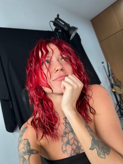 A waist-up shot of a woman with red hair and tattoos on her arms is lying on her side against a black surface, possibly a backdrop, in a room that appears to be a photography studio. A professional camera flash is partially visible in the lower left corner. A cabinet is on the top left wall, while a tripod and a bag are visible on the top of the photo. The location cannot be determined to be in Dalung, Indonesia.