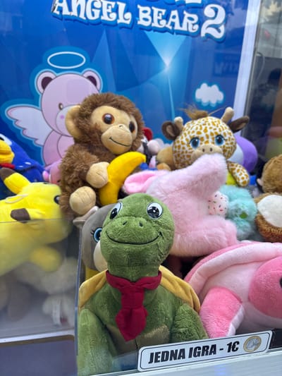 The image shows a close-up view of a claw machine filled with plush toys. The toys include a green turtle with a red scarf, a brown monkey holding a yellow banana, a giraffe, a pink bunny, a yellow Pikachu, and other various stuffed animals. In the background, a blue banner reads "ANGEL BEAR 2". A price tag at the bottom of the machine states "JEDNA IGRA - 1€" which translates to "ONE GAME - 1€". The scene is indoors, likely in a public space given the presence of an arcade game. The lighting is bright, illuminating the colorful toys. There are no people visible in the image. The location is identified as Petrovac centar, Montenegro.