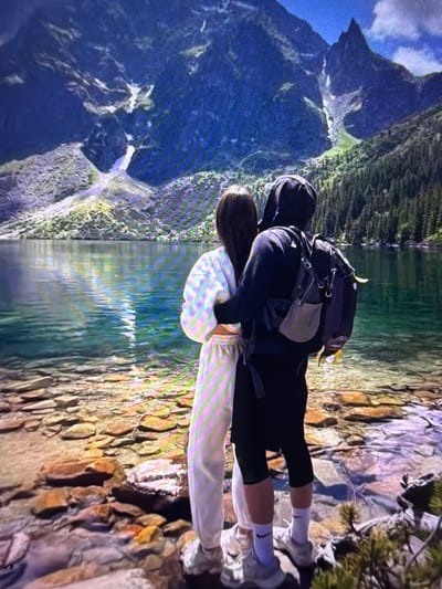 The image displays a couple embracing while standing on a rocky shore overlooking a vast mountain landscape reflected in a body of water. The woman, in the foreground, is wearing a light-colored hooded sweatshirt and off-white sweatpants with a faint pink and yellow striped pattern. The man, behind her, is dressed in a black hooded jacket and dark shorts, with a backpack on his shoulders. Both are wearing athletic shoes with white socks. The scenic backdrop features dramatic, verdant mountains under a bright, cloudy sky, with a clear reflection of the peaks and sky visible in the still water. The lighting suggests daytime. The overall mood is intimate and serene, capturing a moment of connection amidst a grand natural setting. There is no visible text in the image.