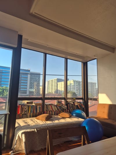 The image shows the interior of a room, likely an apartment or condo, in Pasay, Philippines, based on the view through the windows. The focal point is a large window composed of multiple panes, offering a clear view of the cityscape. Several high-rise buildings are visible, reflecting the clear, blue sky.
Inside the room, to the right of the window, is a bookshelf filled with books of various sizes and colors. Next to the bookshelf is a couch with a throw blanket and pillows, with two large blue exercise balls resting on the seat. Sunlight streams into the room, casting shadows and highlighting textures.
The camera angle is from a high vantage point, looking slightly down and across the room. The overall impression is one of a comfortable, lived-in space with an emphasis on books and a view of the city.