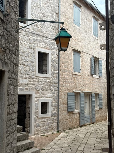The image captures a narrow, cobblestone street in Grad Stari Grad, Croatia, flanked by traditional stone buildings. On the left, a building with rough-hewn stone walls features an open doorway with stone steps leading inside, and a small, square window opening above. To the right, a building with lighter-colored, more uniform stone blocks showcases several windows with pale blue shutters. A vintage lantern with a green hexagonal roof and amber glass panels hangs from an ornate metal bracket extending from the left building, casting a warm glow in the overcast lighting. The street is paved with interlocking stones, and a few sprigs of green grass sprout from the gaps between them. The architecture suggests a historic European setting, characterized by rustic stone construction and functional, aged design elements.