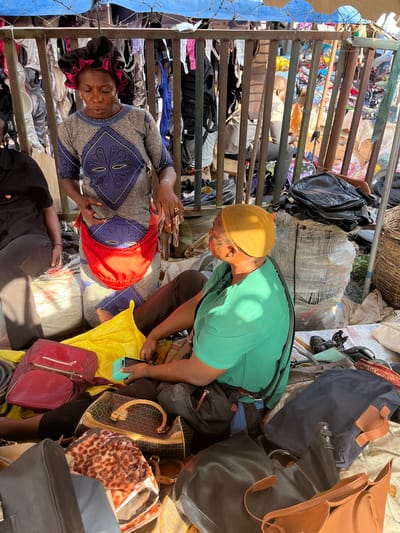 The image depicts a busy marketplace scene in Nyanya, Nigeria, likely during the daytime given the bright sunlight casting shadows. Two individuals are visible, one wearing a green t-shirt and a yellow head covering, bending over and looking at a pile of handbags. The other person, lying down or resting on a bed of clothes and bags, is wearing a blue and white patterned dress and a dark head covering. They are also holding a smartphone.
The scene is characterized by an abundance of items, primarily clothing and a large collection of handbags of various styles, colors, and brands. Some of these include a large dark blue tote bag, a brown leather bag, a black backpack, a red backpack, and a patterned handbag with a leopard print. The setting appears to be an outdoor market or a storage area for goods, with metal shelving and piles of merchandise. There are also large woven baskets and bundles of fabric or clothing visible in the background.
The overall impression is one of commerce and activity, with a focus on the sale or display of goods. The individuals' postures suggest they are either involved in sorting, selling, or perhaps purchasing these items. The presence of smartphones indicates modern elements within the traditional market setting. The lighting suggests it is a bright day. No specific text is legible in the image.