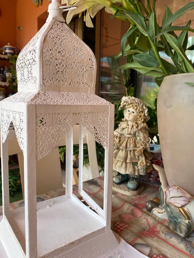 This image depicts a close-up, rotated view of decorative items on a patterned surface. The primary subjects are a white, ornate metal lantern with intricate cut-out patterns and a small, detailed figurine of a young girl. The figurine, dressed in a ruffled dress and bonnet, appears to be holding a basket. Behind these objects, a large, light-colored ceramic pot with greenery spilling out is visible. The background suggests an indoor setting with a table covered by a fabric featuring a red and beige plaid pattern interspersed with small red hearts and red ribbon motifs. The lighting is soft, indicating it could be daytime with diffused light or indoor lighting. The overall mood is quiet and decorative. No text is visible in the image. The location context is Resinera-voladilla, Spain.