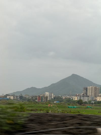 The image captures a view of the city of Vasai-Virar, India, under a cloudy sky. A prominent, verdant mountain range forms the backdrop, its slopes softened by the overcast weather. In the midground, a cluster of buildings, ranging from residential complexes to smaller structures, is visible, nestled against the base of the hills. These buildings display a variety of architectural styles and colors. In the foreground, a lush green field stretches out, dotted with some agricultural elements like what appear to be small, temporary shelters covered with green tarpaulin, and what might be scaffolding or supports for some kind of structure. Two small, pink objects, possibly mannequins or decorative items, are positioned in the field, adding a touch of color to the otherwise natural and urban landscape. The foreground also shows a blurred section that suggests the image was taken from a moving vehicle, with a dark, textured element in the immediate foreground.