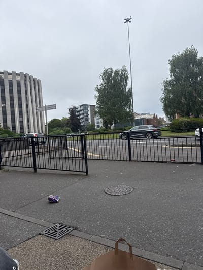 A dull, overcast day is depicted in this medium shot, taken from a low angle on a pedestrian walkway in Poole, United Kingdom. A black metal fence runs horizontally across the foreground, dividing the walkway from a road. On the walkway, a crumpled piece of purple and silver packaging lies on the pavement, near a square drain cover.
Beyond the fence, two lanes of asphalt road are visible, with a white line marking the center. A dark grey Audi SUV is driving in the right lane, while a white car is visible further down the road in the left lane. Several trees, some with dark foliage and others with lighter green leaves, line the far side of the road. Behind the trees, a modern apartment building with balconies and a strip of windows can be seen, along with a taller, grey building with vertical concrete fins on its facade, which is characteristic of some office blocks. In the distance, a tall, slender lamppost with multiple lights stands against the sky. The overall scene suggests an urban environment, likely a commercial or residential area within Poole.