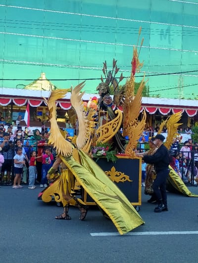 A cultural parade is depicted on a paved street in the city of Madiun, Indonesia. A prominent green reflective glass building, labeled "M. CAFE & RESTO" in black text, is visible in the background. To the left of this building, a structure with a white balustrade and red and white striped decorative fabric, along with an Indonesian flag, is present.

The central subject is a large, elaborate parade float, predominantly black and gold. The float features multiple sets of large, ornate golden wings and gilded scrollwork on its black base. A figure in an elaborate dark costume, complete with a helmet-like headpiece adorned with vertical spires and golden shoulder pieces, stands on the central platform of the float. Small, colorful figures, including a yellow cartoon-like character, are also visible on the float.

A participant in a golden costume, featuring large golden wings and a long golden train, walks in front of the float, seemingly attached to or pulling it. This individual wears dark sandals. On the right side of the float, a man wearing a black baseball cap, black long-sleeved shirt, black pants, and black shoes is actively pushing the float. Another individual, dressed in a patterned garment, stands alongside him near the float.

A crowd of spectators, comprising adults and children, lines both sides of the street behind a dark barrier. They observe the parade, with several individuals holding up mobile phones or cameras. The street surface is dark asphalt with a single white painted line. Overhead utility wires are visible against a bright, clear sky. The scene is illuminated by natural daylight.